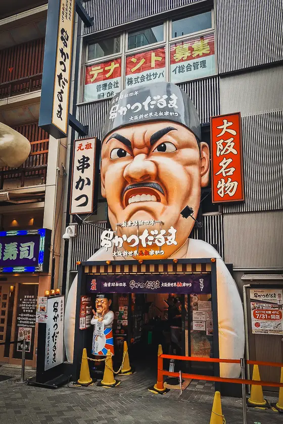 Iconische ingang van Kushikatsu Daruma in Shinsekai, Osaka, met het grote karikatuurhoofd boven de gevel.